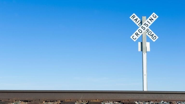 Under the horizon of a pale blue sky is a rail crossing sign stood on a pole and shaped like an x representing our report on rail regulation in Washington.