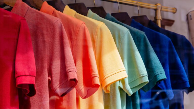 Celebrating June Pride Month at Cowen with Cowen OUTPerformers and Macy's. The image blends retail with pride with polo shirts aligned in succession of the colors of the rainbow pride flag are hung on a gold dresser in a bright sun-lit room.
