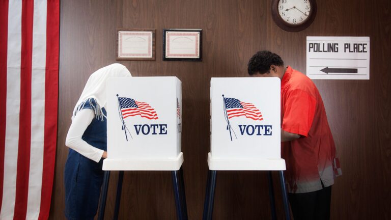 Two people about to vote in U.S midterm elections. Anonymity is secured as the two people stood across from each other have their heads buried in a voting booth ordained in American flags with the slogan "vote".