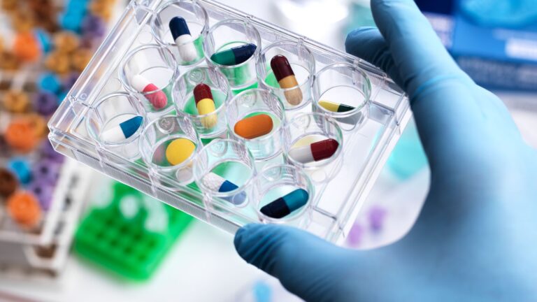 Man in a lab coat and gloves is holding a petri dish fit for several different shapes of pills. Representative of big pharma versus smaller U.S based biotechs.