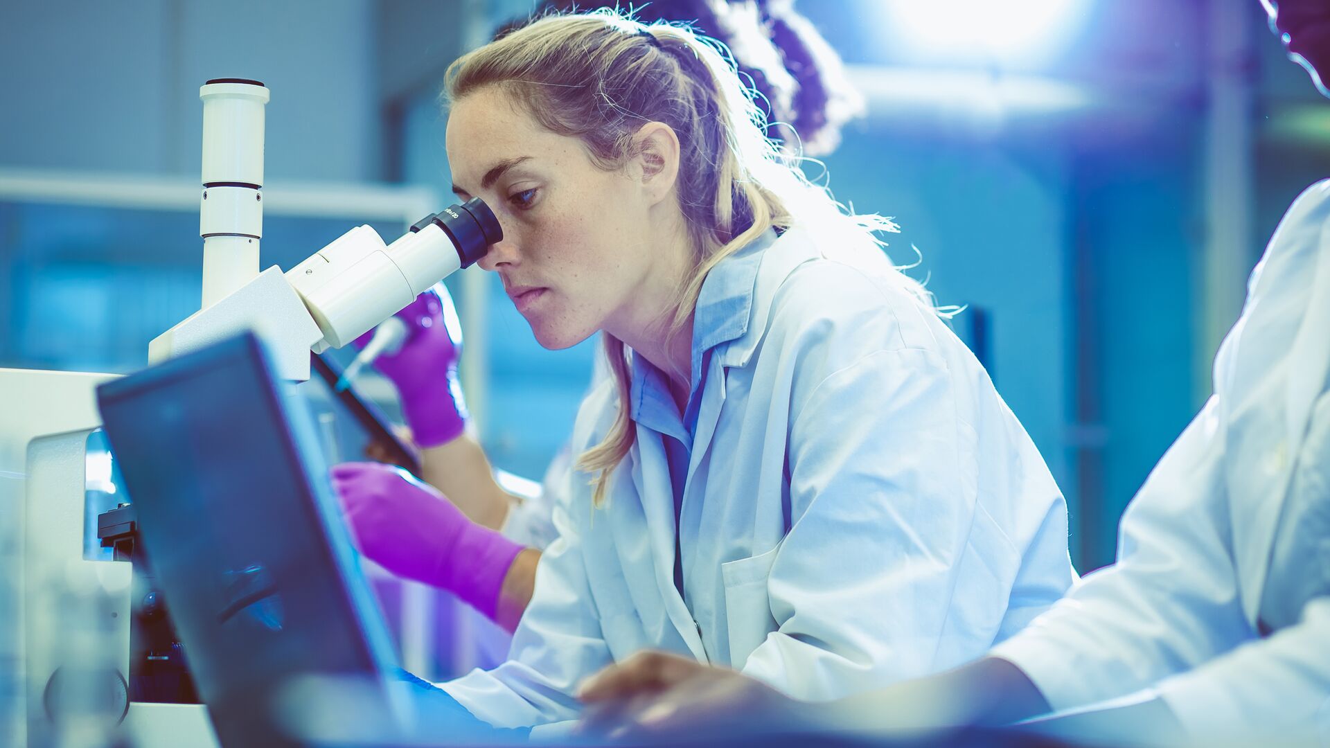 Female lab technician looking under a microscope at cancer cells. Representative of Liquid Biopsies.