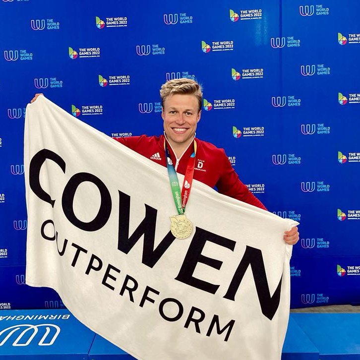 Malte Strieger holding a "Cowen Outperform" flag while sporting a gold medal around the neck, smiling triumphantly at the camera.