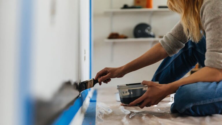 Woman painting a wall a dark color, with blue painter's tape set up to create lines