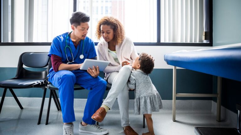 A medical professional sitting down with a woman who has a child. The medical professional is showing the woman something on a tablet device.