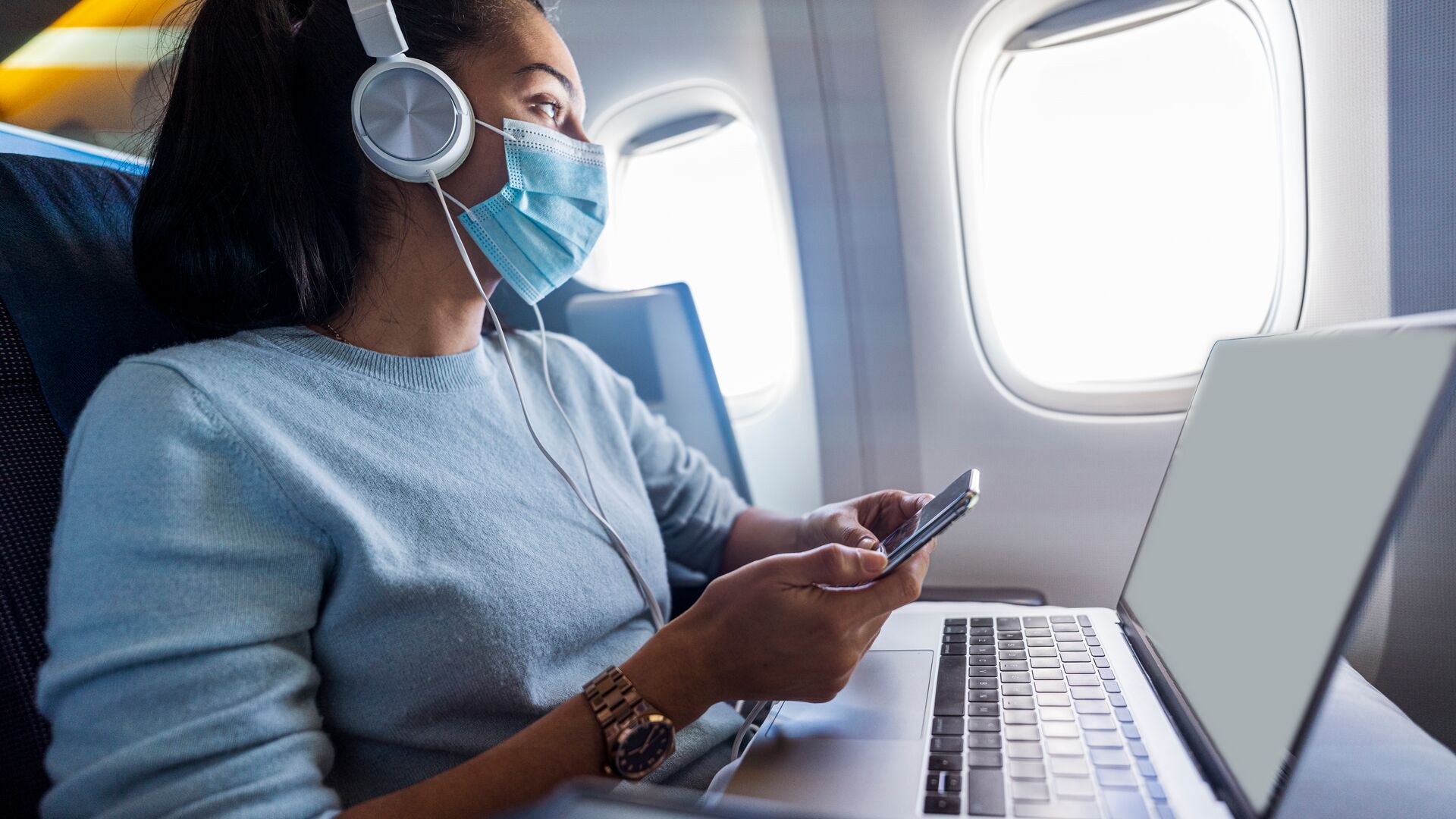 Women waiting for plane at gate on her phone with a mask on to prevent the spread of Covid-19 while a plane takes off behind her. Representing the post-pandemic environment for airlines.