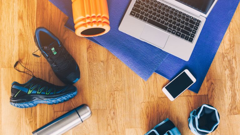 Running shoes, yoga matt, water, and laptop are laid across a wooden floor in someone's home representing fitness and wellness in the consumer space.