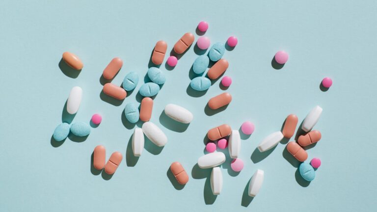 A variety of pharmaceuticals laid on top of a light blue background. Laid on top are a variety of pills of various colors.