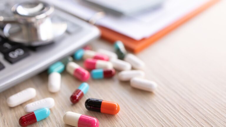 A variety of pharmaceuticals laid on top of a desk next to a calculator, representing our drug pricing outlook. Laid on top are a variety of pills of various colors.