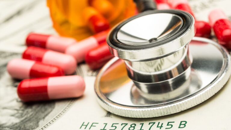 Pills laid on top of a twenty dollar bill next to a orange pill bottle and a stethoscope, as if it were measuring the health of the money and pills. Representing drug pricing in the U.S.