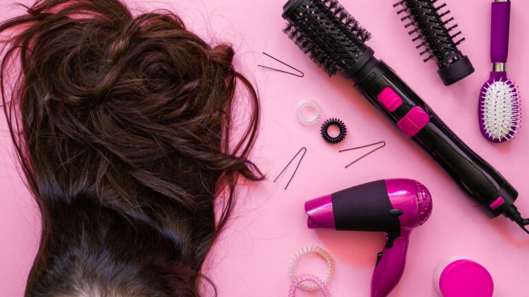 Laid on a pink table is a brown haired wig blow drier and other hair products and tools representing a recent TD Cowen retail visionaries podcast with Sally Beauty.