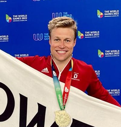 Malte Strieger holding a "Cowen" flag while sporting a gold medal around the neck, smiling triumphantly at the camera.