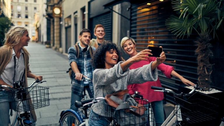 Young shoppers on bicycles posing for a group selfie during the day in an alley way in the city.