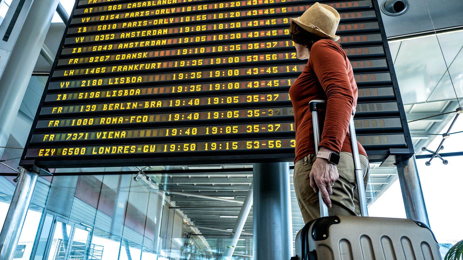 Female traveler with luggage in tow is looking at flight information which details flights travelling around the world.