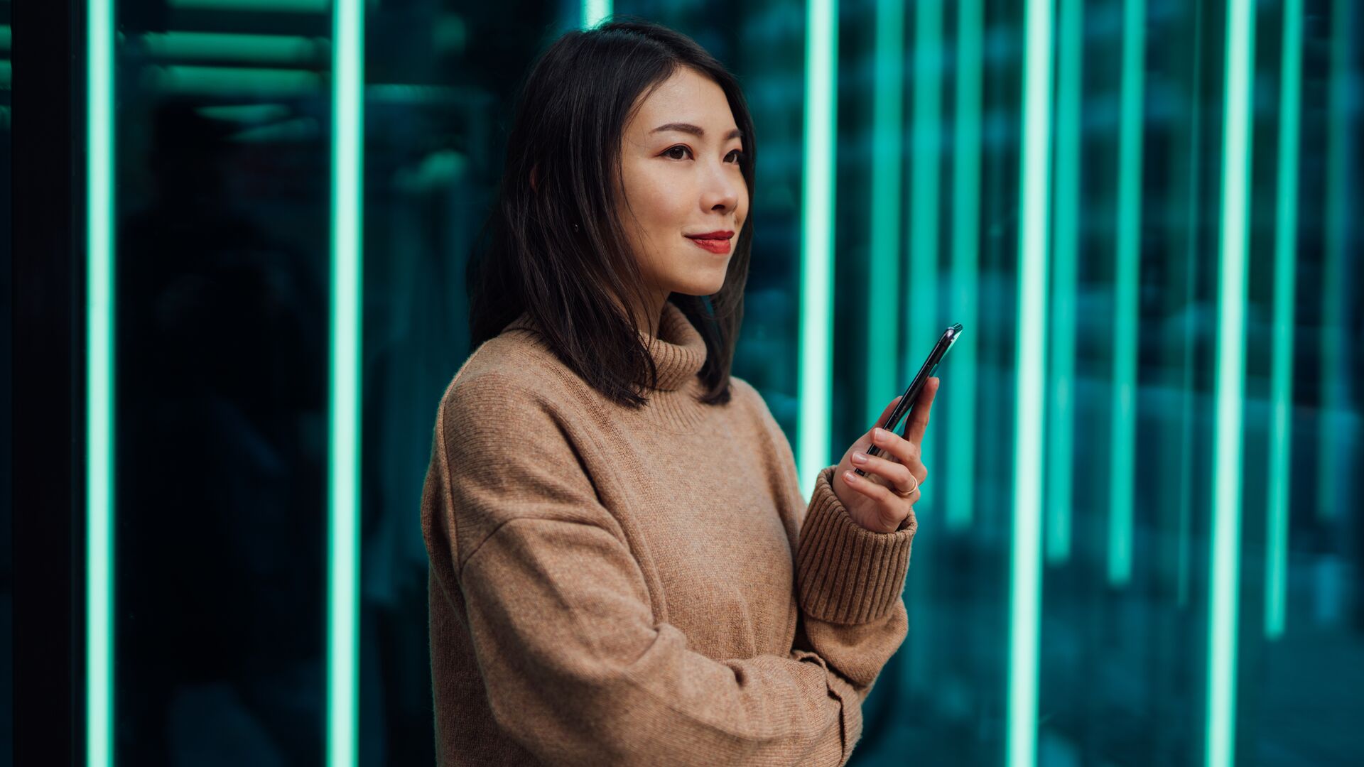 Woman using a mobile phone in front of a neon blue-green lit space.