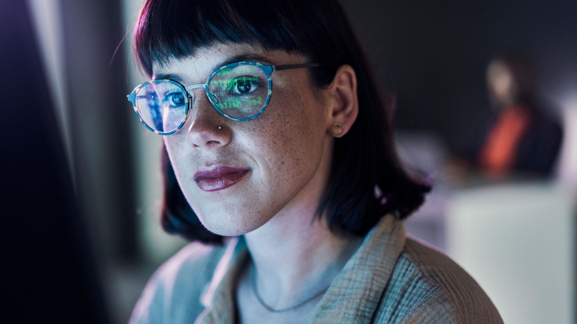 One's and zeros in green reflect against a woman's glasses who is looking intently in front of a screen with another woman in a similar position in the background out of focus.