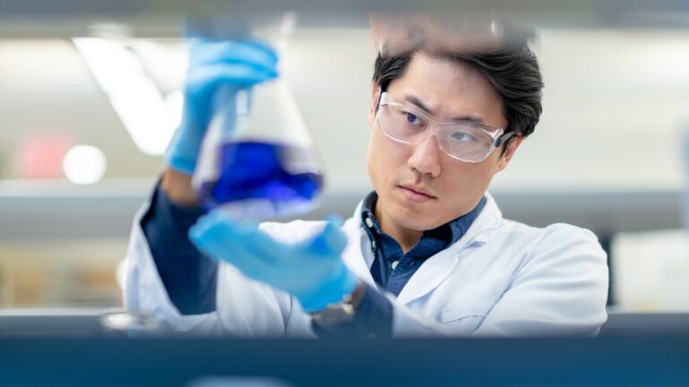 Biochemist in lab wearing lab coat. blue gloves, and goggles is looking at a blue liquid solution in a glass flask.