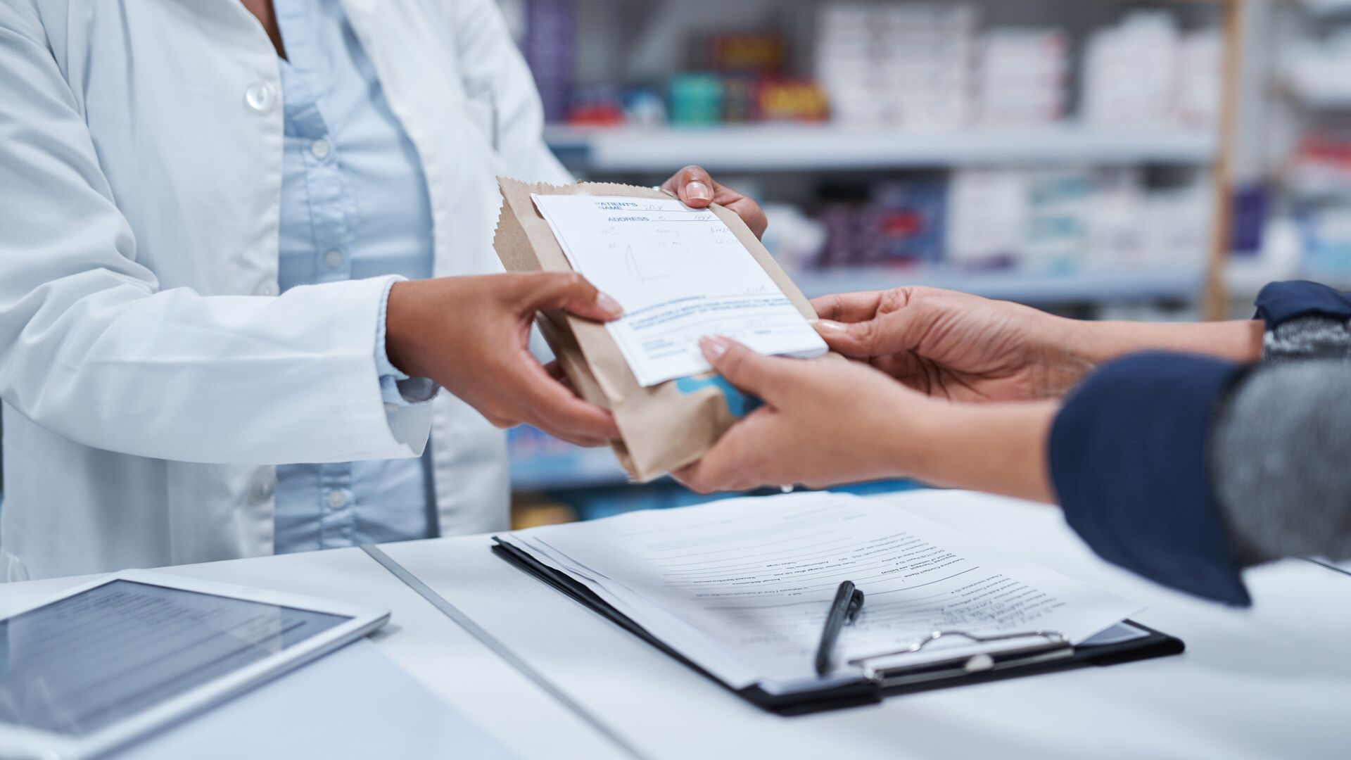 A pharmacist assists a customer by handing them their medication in a brown paper bag across the counter at a pharmacy.