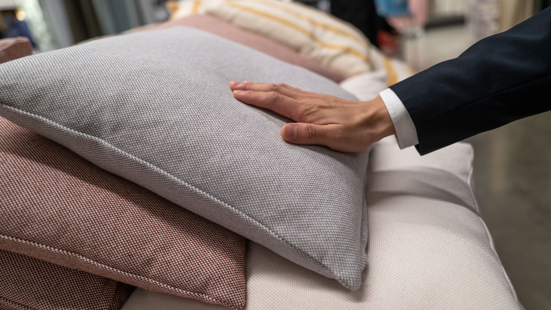 A hand of an adult male in a suit jacket touching a grey pillow.