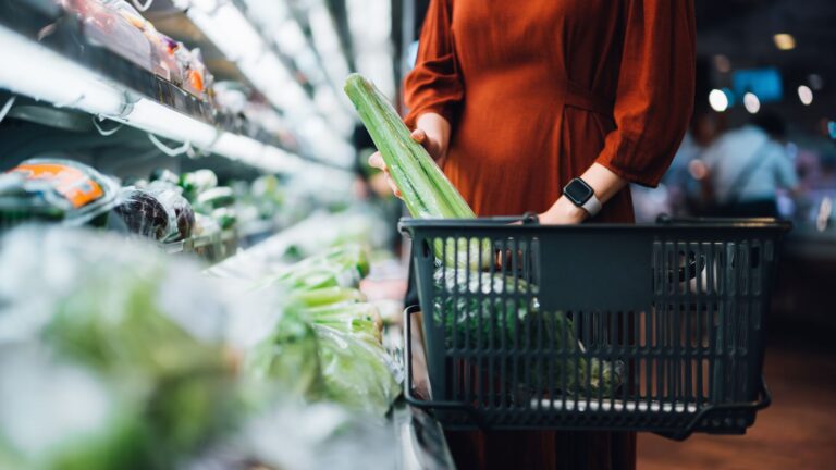 A woman grocery shopping with a huge stalk of celery visible in her hand held shopping cart.